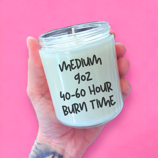 A hand holding a candle labeled "MEDIUM 90Z 40-60 Hour Burn Time" against a pink background.