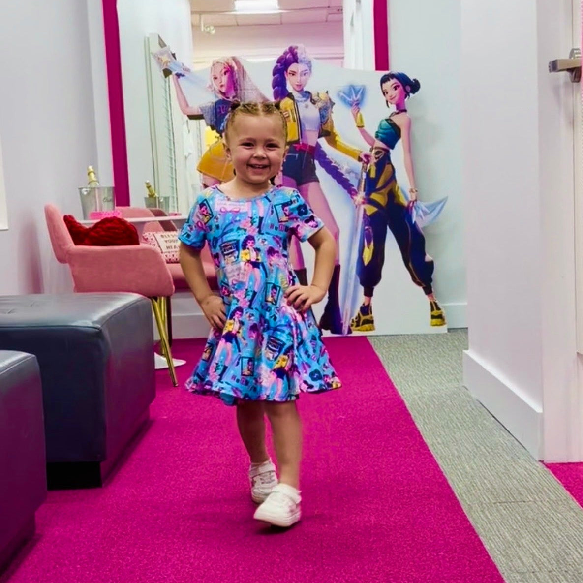 A young girl stands in a hallway wearing a colorful blue patterned dress, with a large pink-framed mirror behind her showing cartoon characters and a pink chair to the left.