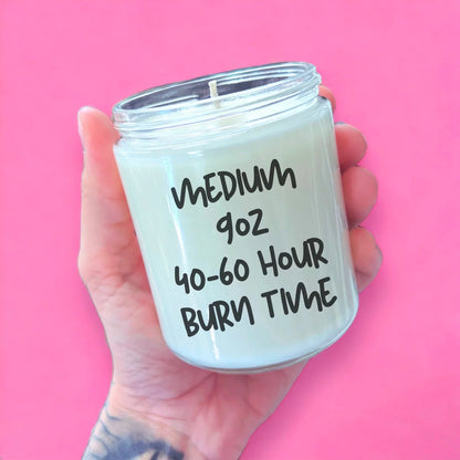 A hand holding a candle labeled "MEDIUM 90Z 40-60 Hour Burn Time" against a pink background.